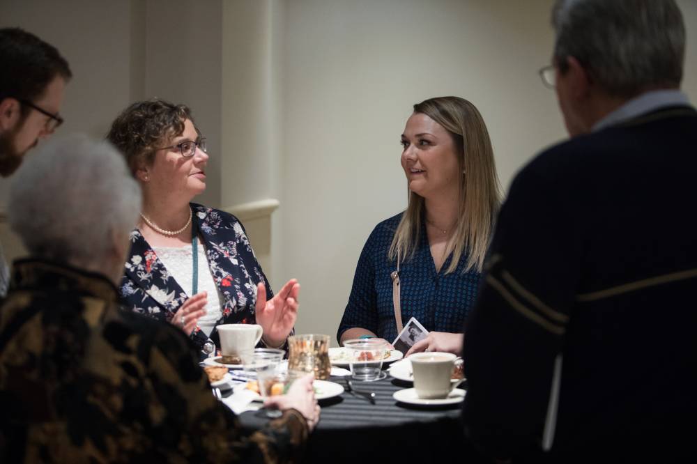 Two women talking with a group of people at a table before the Fall 2019 Graduate Student Celebration